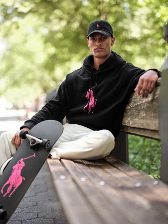 Young man sitting on a park bench holding a black skateboard wearing a black hoodie and cap.