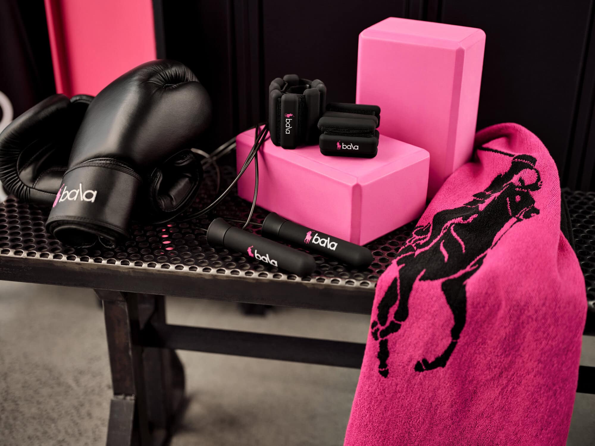 Black boxing gloves, jump rope, wrist weights, pink yoga blocks and a pink towel with black branding arranged on a dark mesh bench.