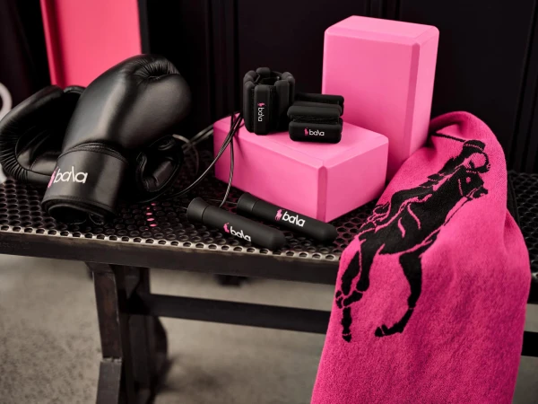 Black boxing gloves, jump rope, wrist weights, pink yoga blocks and a pink towel with black branding arranged on a dark mesh bench.