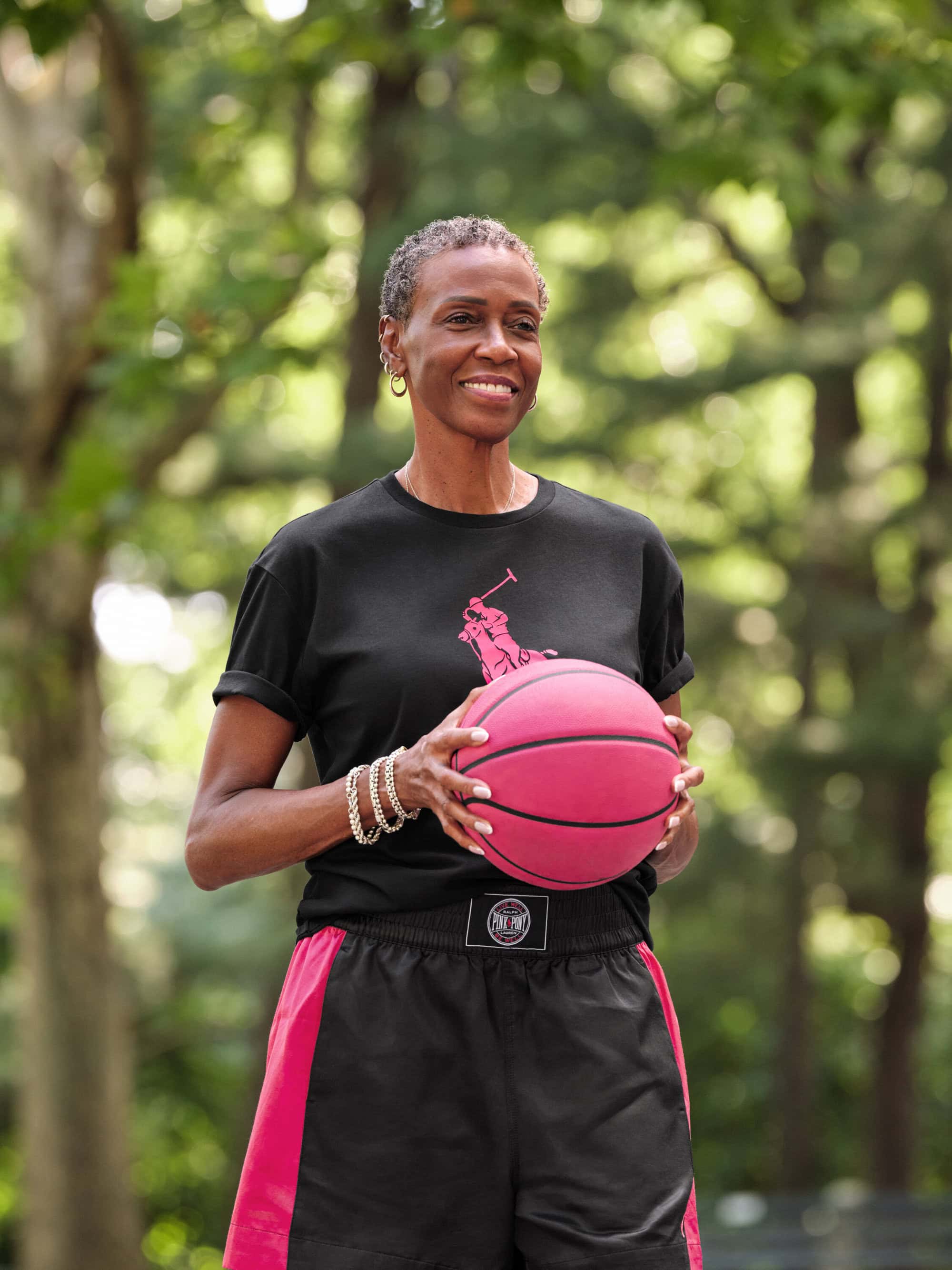 An older Black woman smiling while holding a pink basketball, wearing a black t-shirt and shorts in a park.
