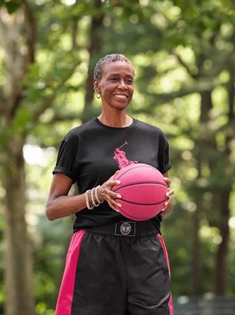 An older Black woman smiling while holding a pink basketball, wearing a black t-shirt and shorts in a park.
