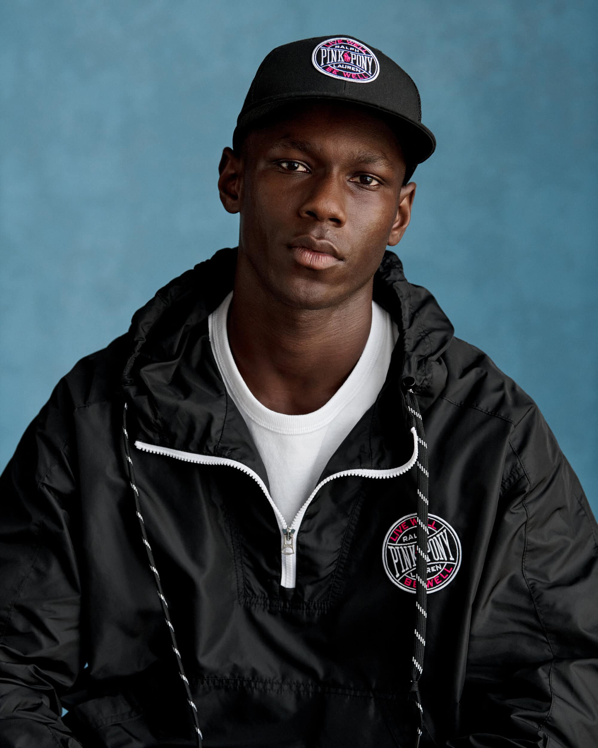 Close-up portrait of a Black man wearing a black anorak hoodie, white t-shirt, and black baseball cap against a blue background.