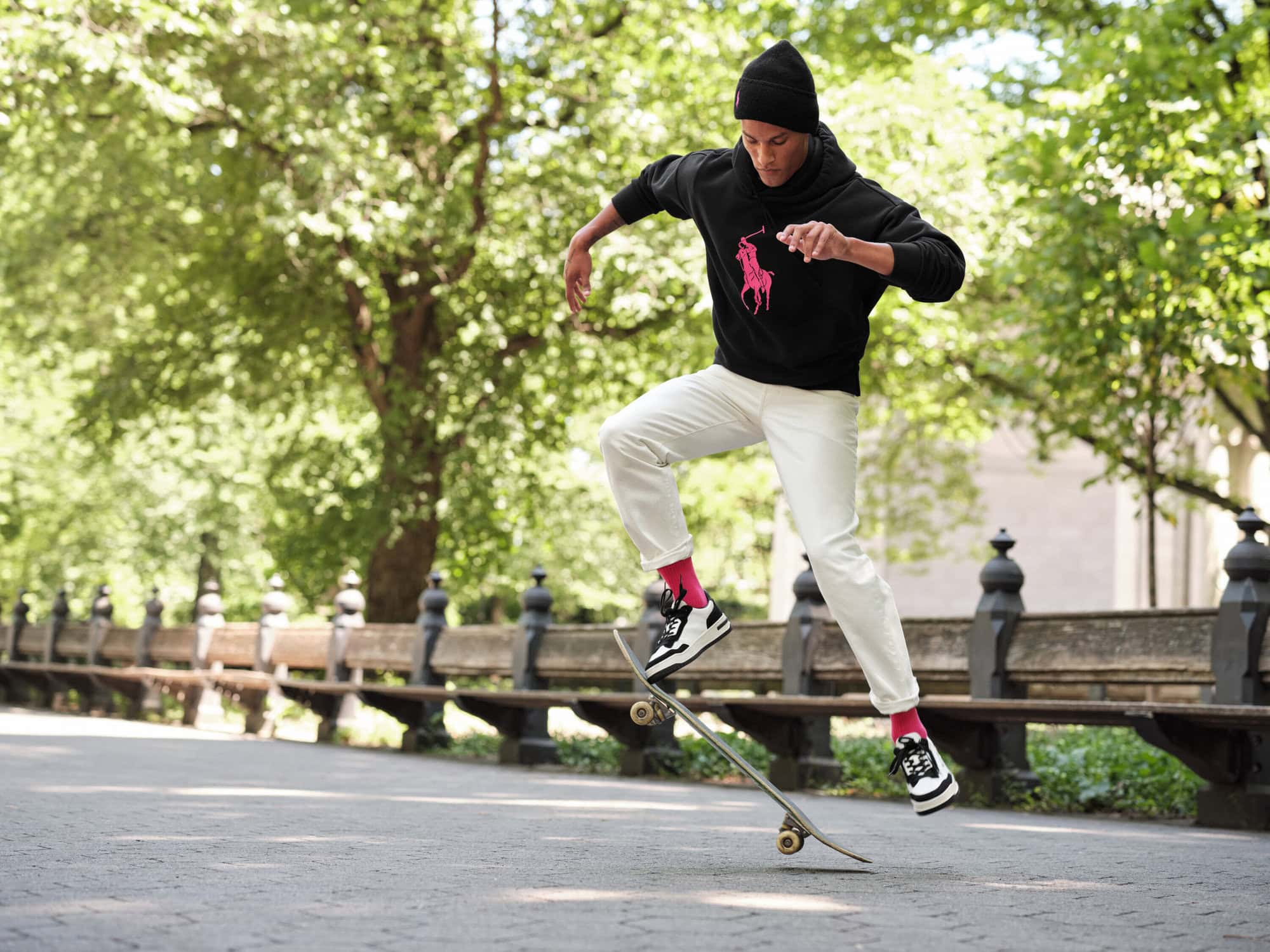 Man wearing a black hoodie with a pink logo and white trousers jumping on a skateboard on a park path