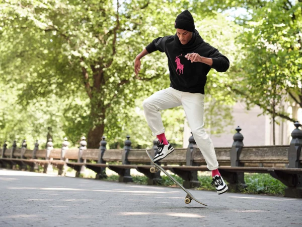 Man wearing a black hoodie with a pink logo and white trousers jumping on a skateboard on a park path
