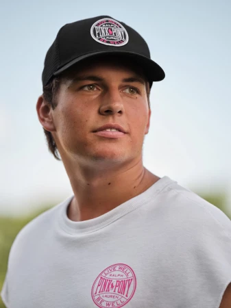 Man wearing a black baseball cap with pink branding and a white t-shirt against a soft-focus outdoor background.