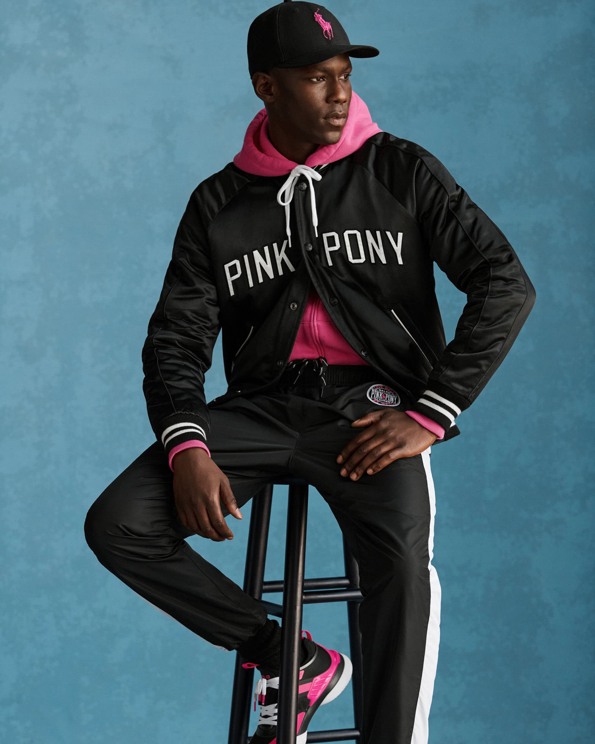 Man wearing a black satin jacket with pink hoodie sitting on a stool against a textured blue background.