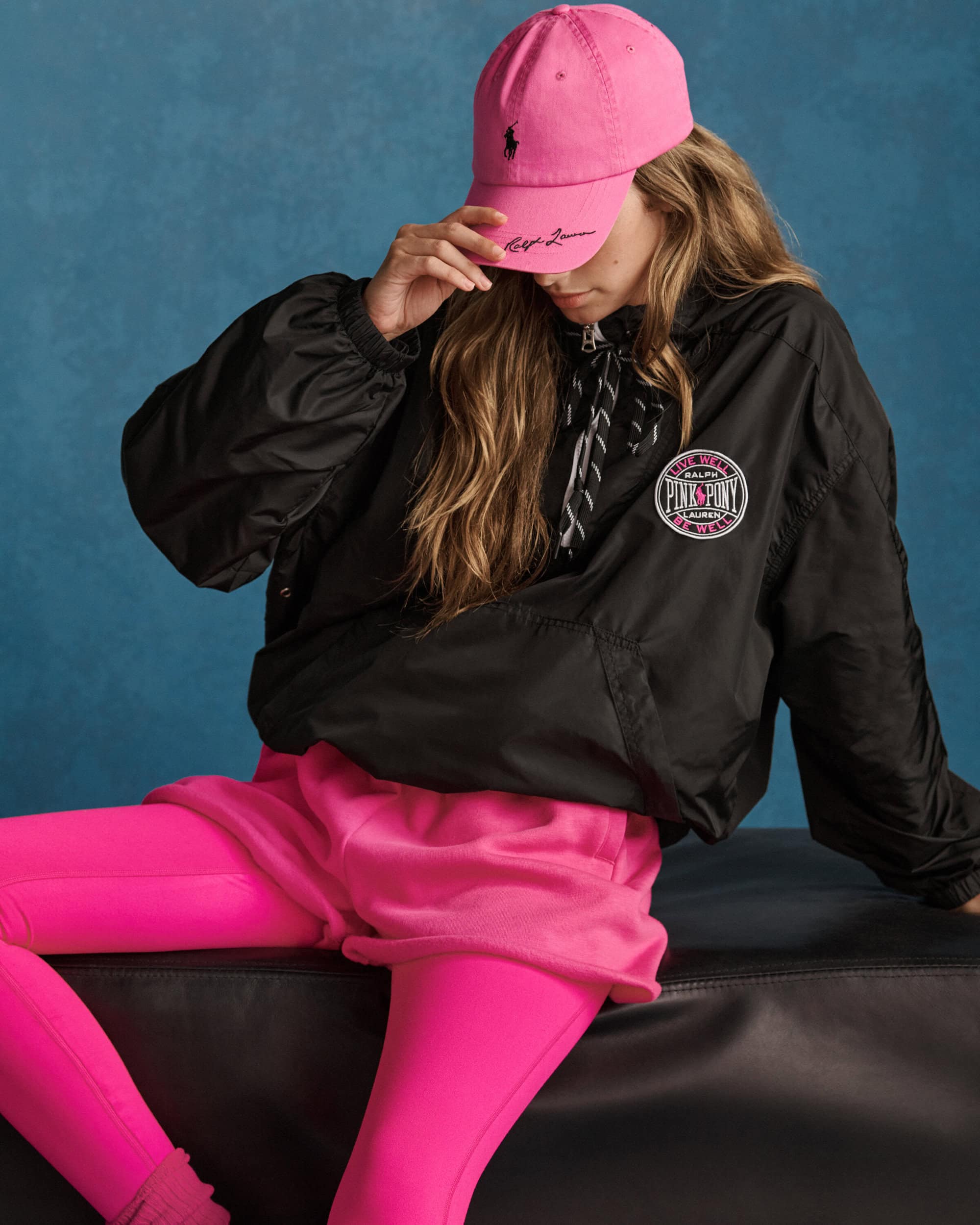 Model wearing a pink baseball cap and black jacket sitting against a textured blue background for Ralph Lauren Pink Pony campaign