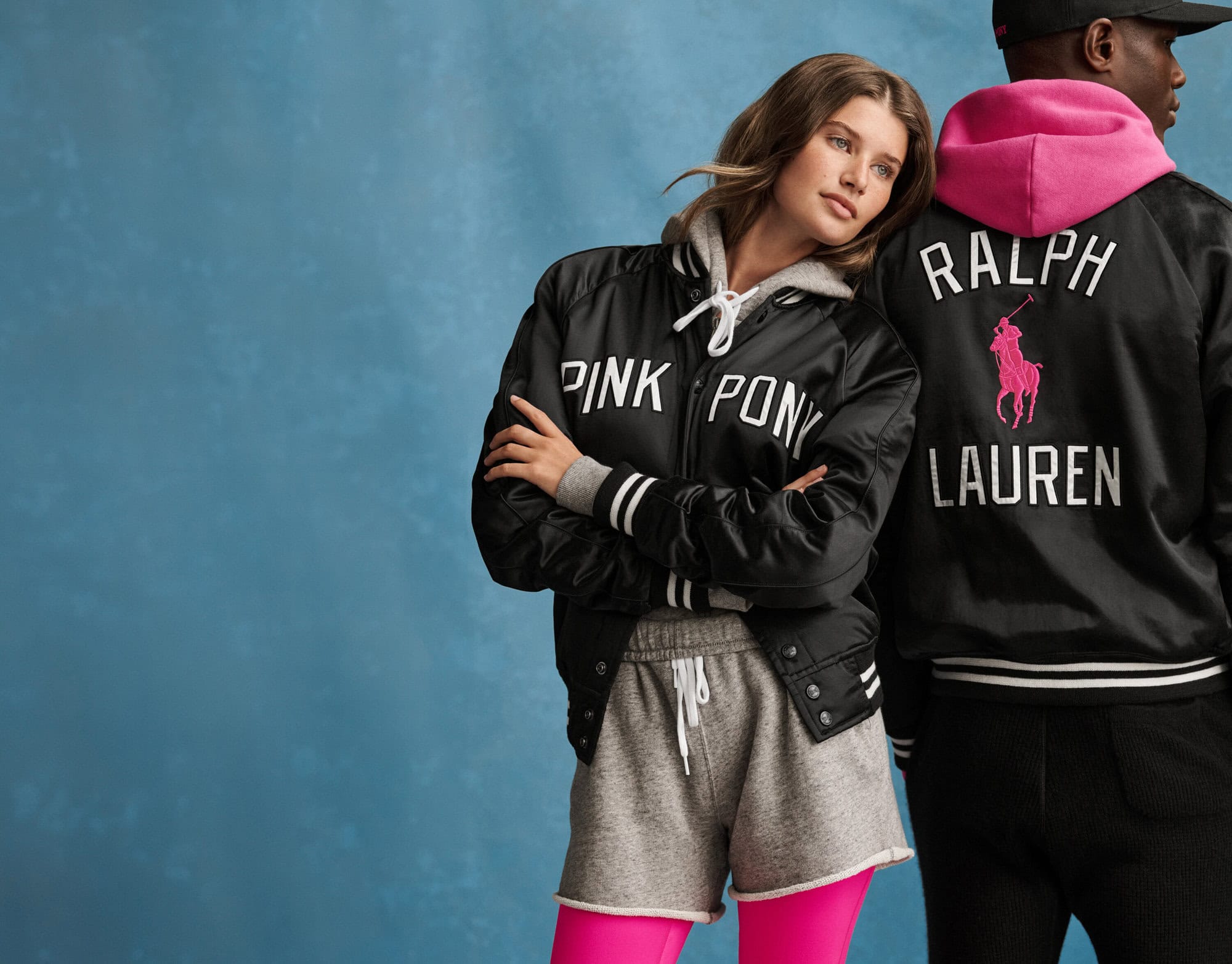 Two models wearing black satin jackets with pink lettering and grey athletic apparel against a textured blue backdrop