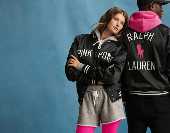 Two models wearing black satin jackets with pink lettering and grey athletic apparel against a textured blue backdrop