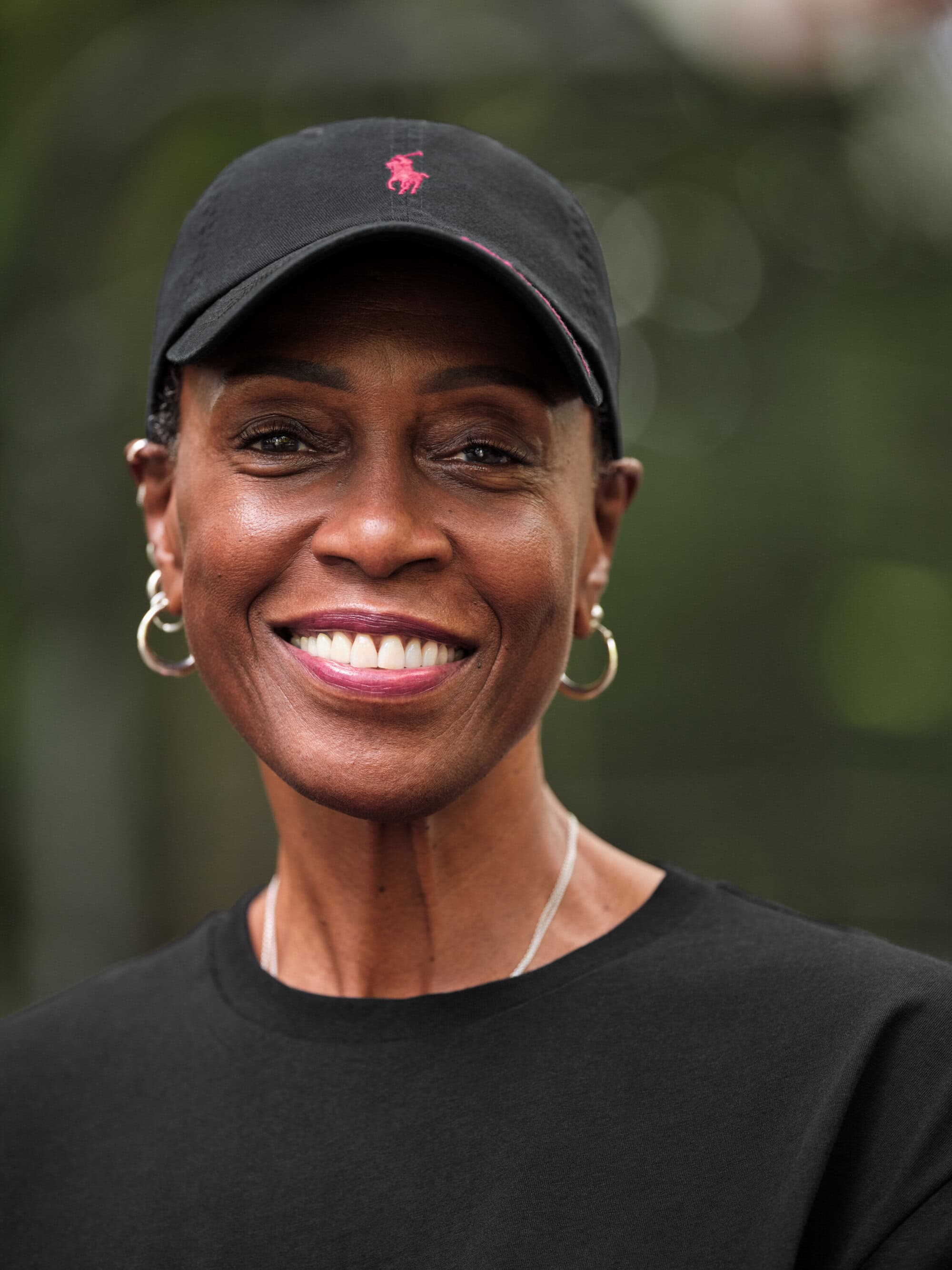 Portrait of a Black woman smiling while wearing a black baseball cap with a pink logo and a black t-shirt
