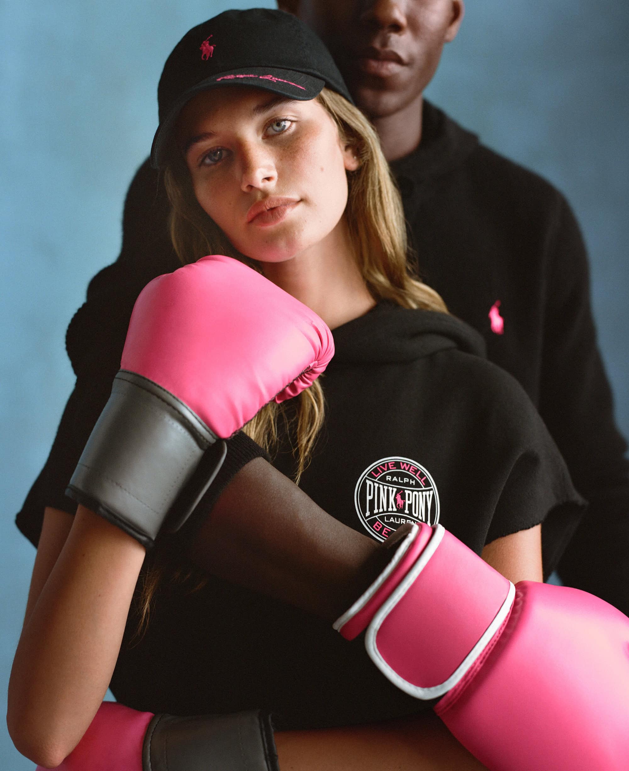 Two models wearing black hoodies and pink boxing gloves posing against a textured blue studio background for Ralph Lauren.