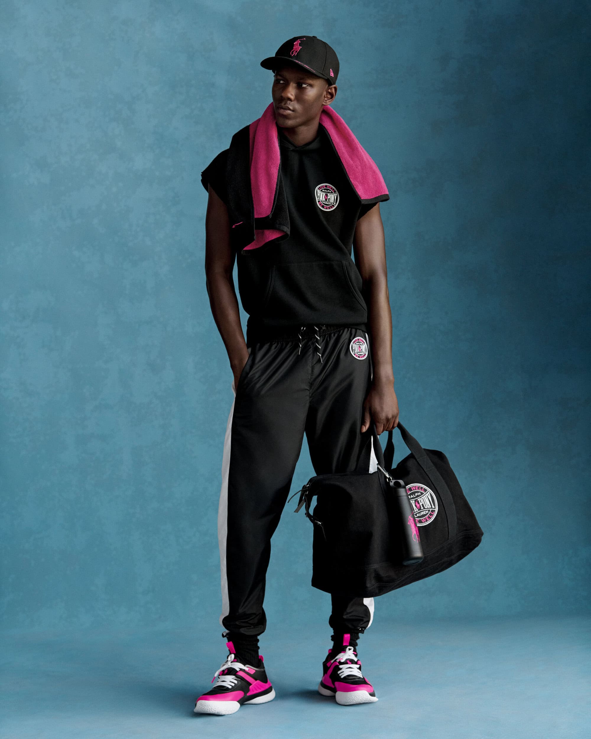 Man wearing a black cap, hoodie, and sweatpants with a bright pink towel around his neck holding a black duffel bag against a blue backdrop