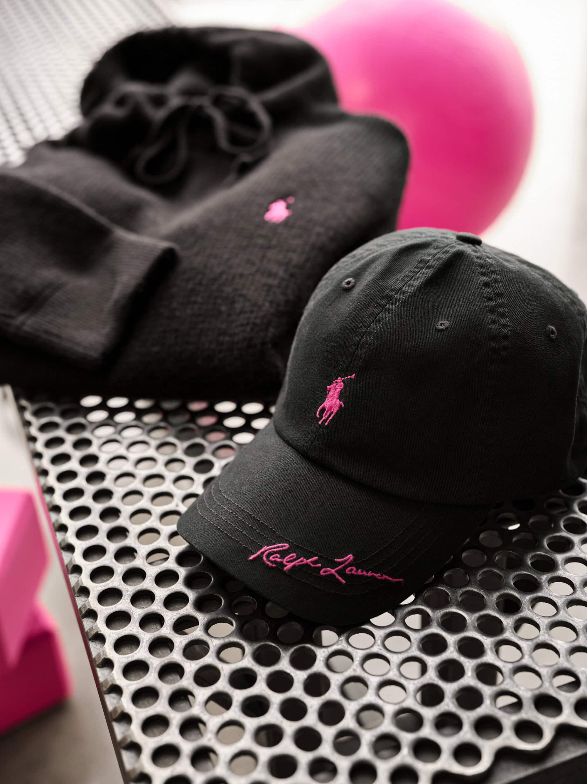 Black Ralph Lauren cap and hoodie with pink embroidered logos on a textured metal surface.