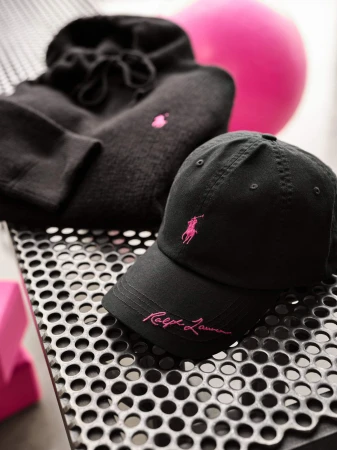 Black Ralph Lauren cap and hoodie with pink embroidered logos on a textured metal surface.