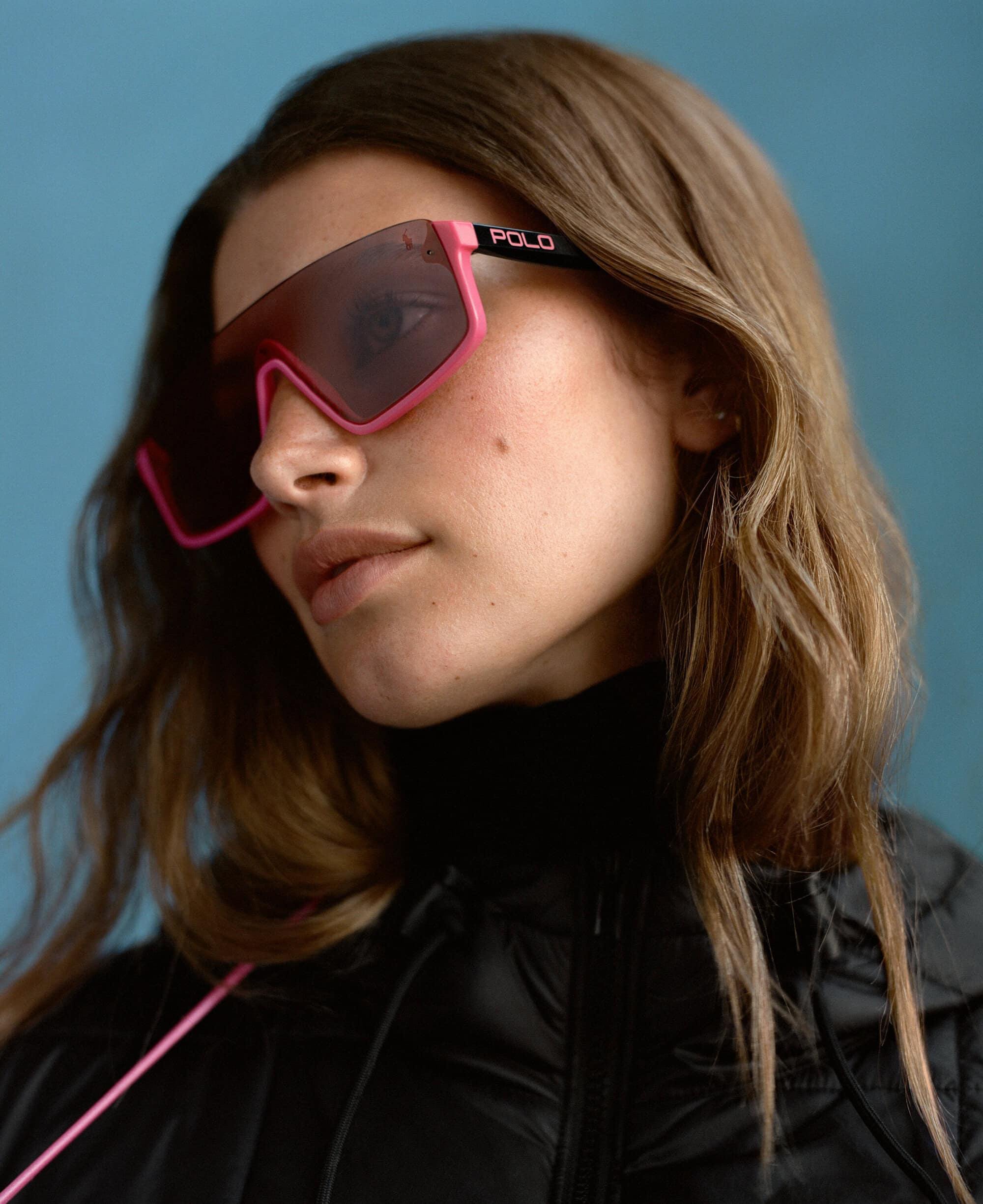 Close-up portrait of a model wearing bright pink oversized sunglasses and a black puffer jacket against a blue textured backdrop.