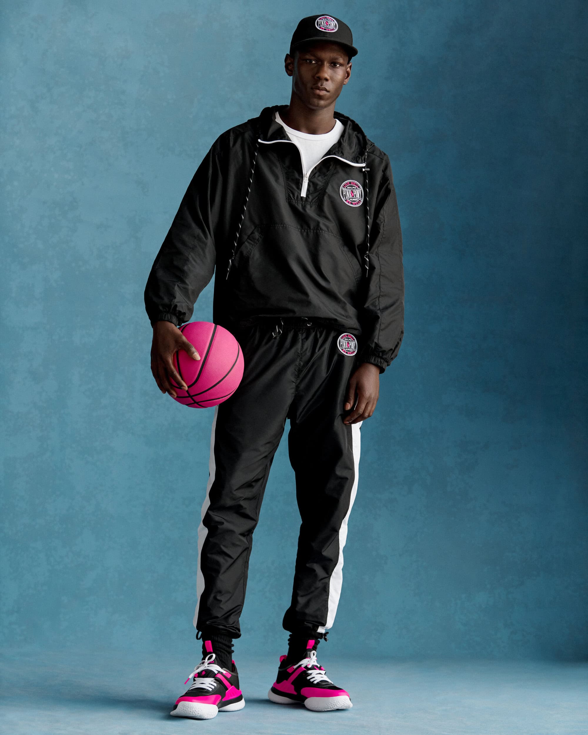 Man wearing a black windbreaker holding a bright pink basketball against a textured blue backdrop