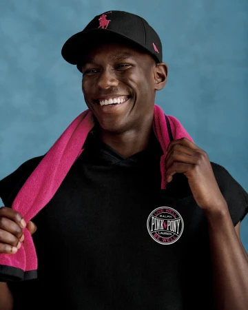 Black man smiling wearing a black baseball cap and black polo shirt with a vibrant pink towel draped over his shoulders.