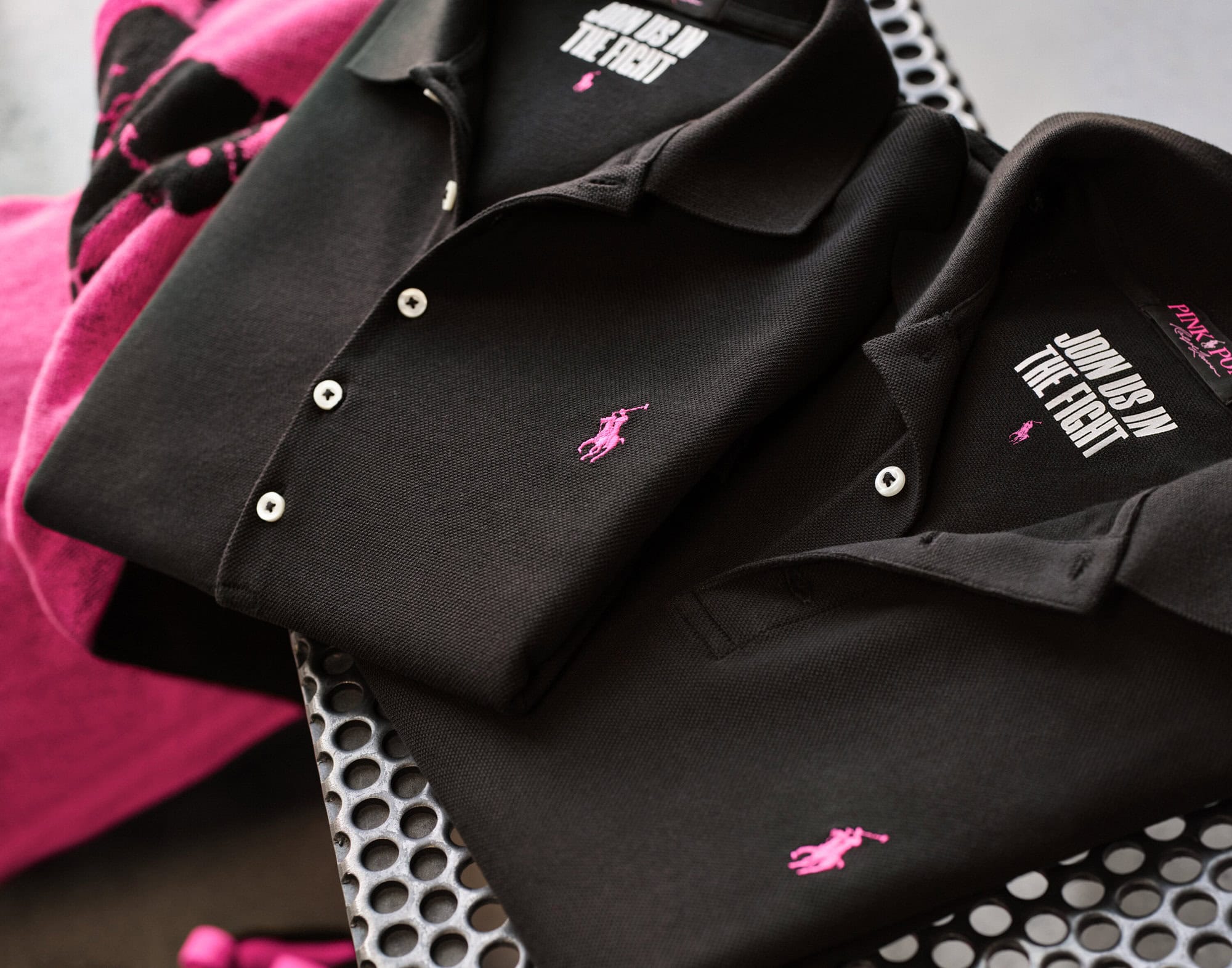 Two black polo shirts with pink embroidered logos stacked on a perforated metal surface