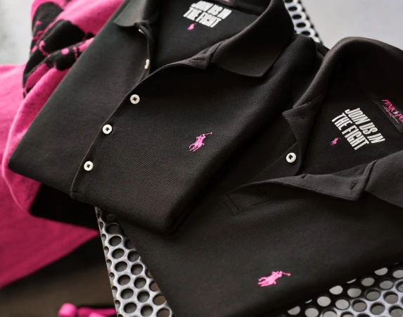 Two black polo shirts with pink embroidered logos stacked on a perforated metal surface
