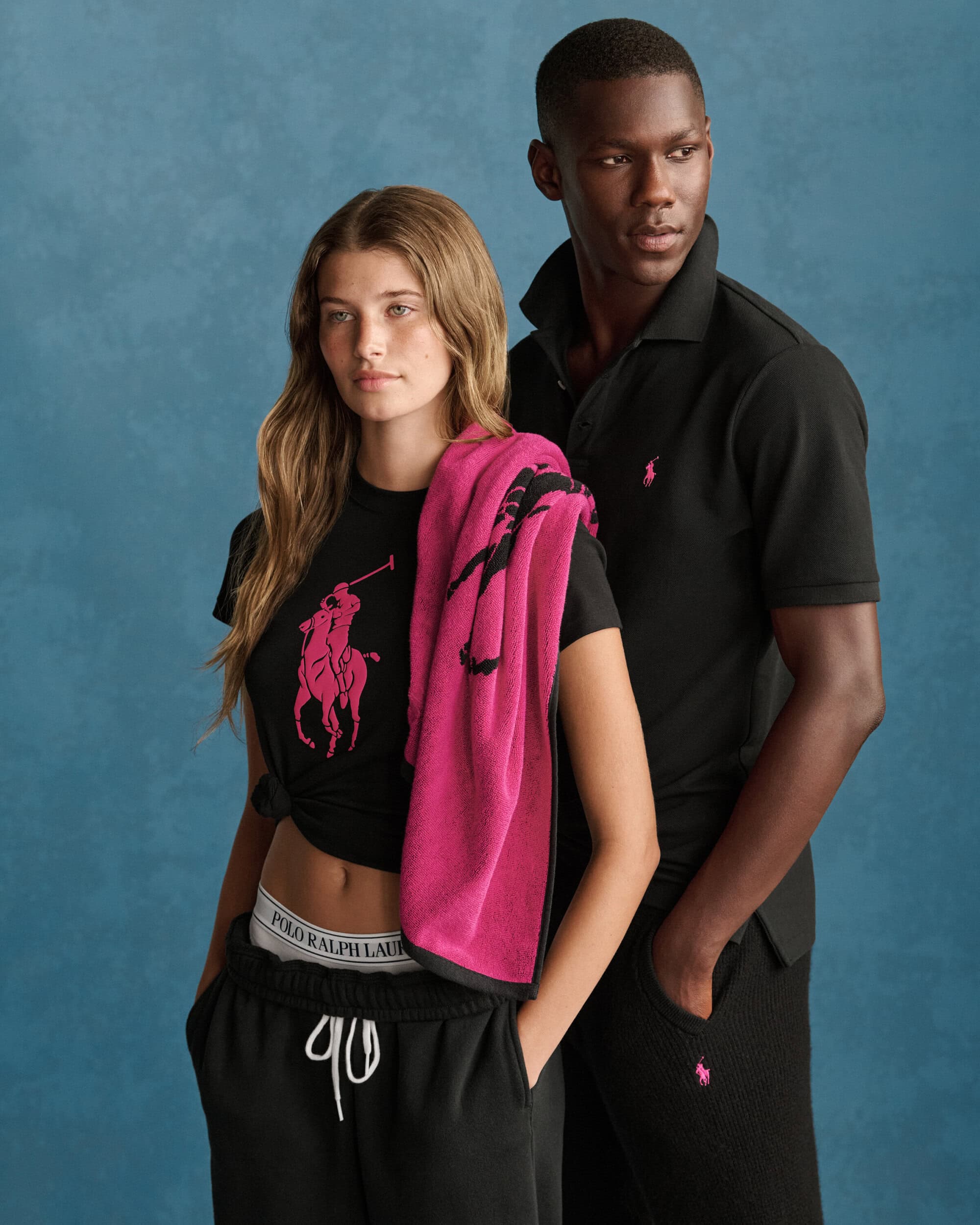 Two models wearing black polo shirts and matching apparel with pink Ralph Lauren branding against a textured blue background