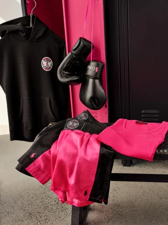 Black hoodie, pink and black boxing shorts, boxing gloves, and a pink tank top displayed in front of a pink and black locker.
