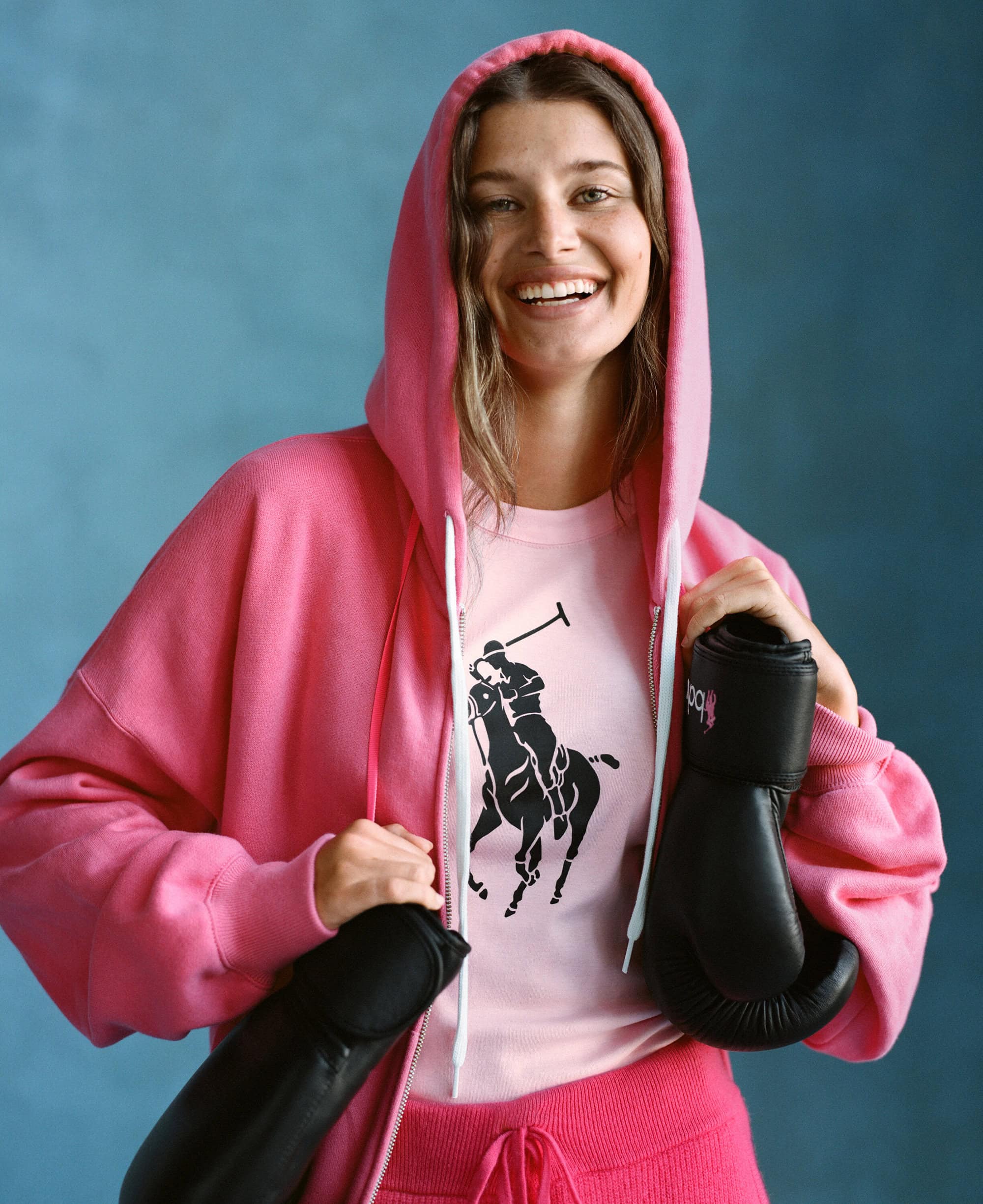 Smiling woman in a pink zip-up hoodie holding black boxing gloves for the Ralph Lauren Pink Pony Fall 2024 campaign.