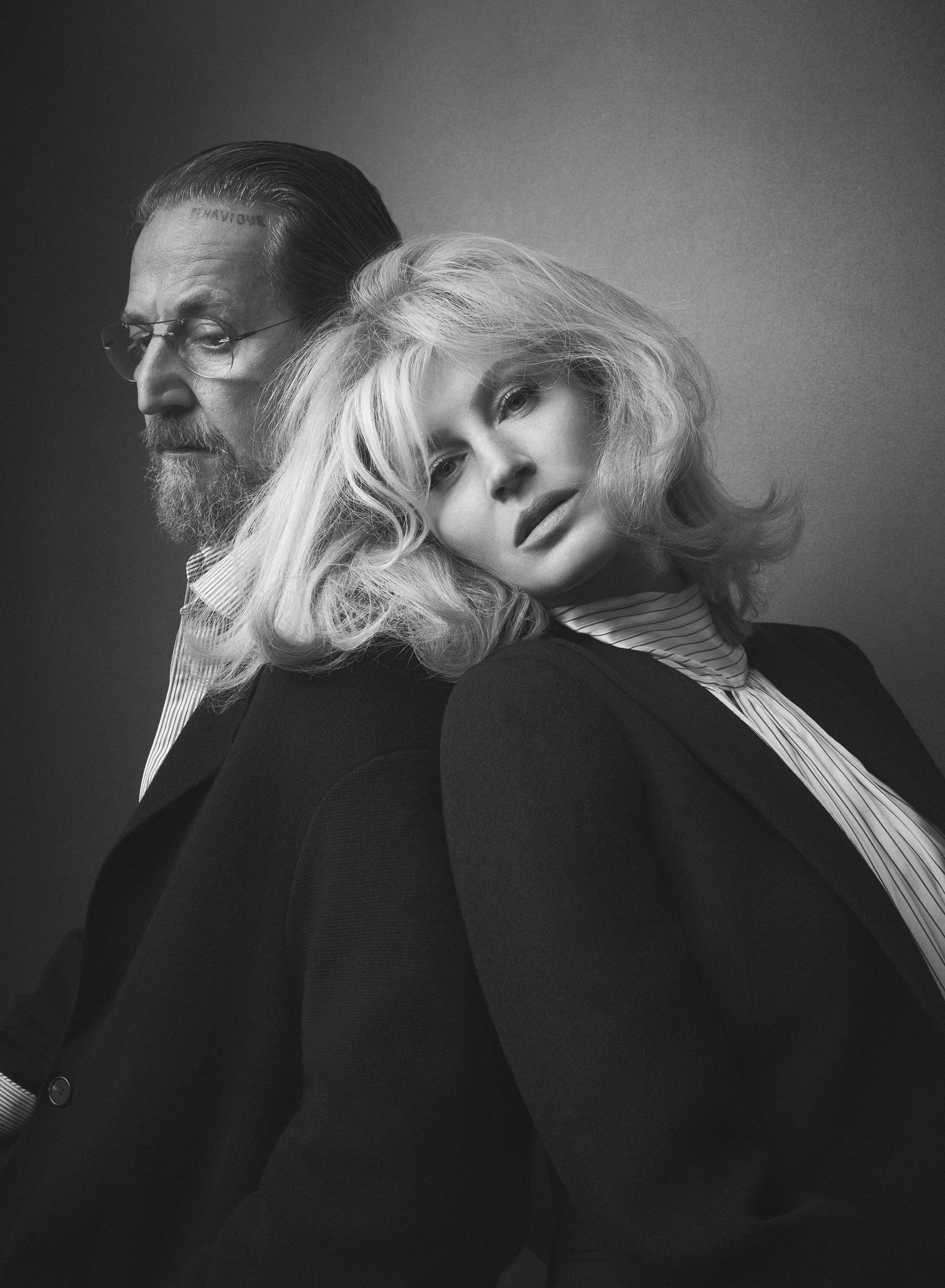 Black and white portrait of a man and blonde woman leaning back to back wearing dark blazers against a neutral studio background