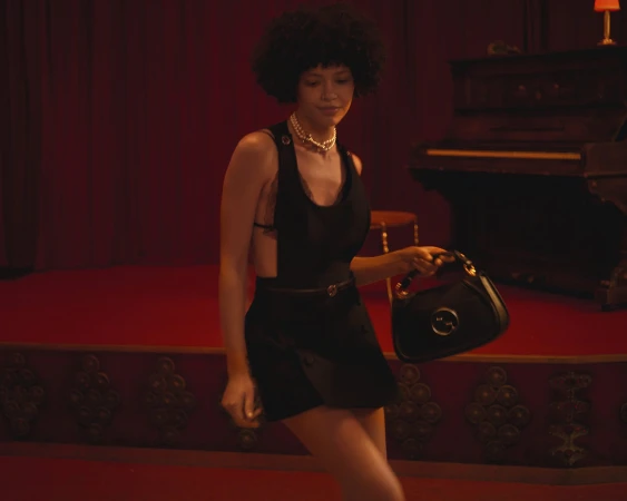 Model with dark afro hair wearing a black dress and pearl necklace holding a black handbag inside a red-lit interior.