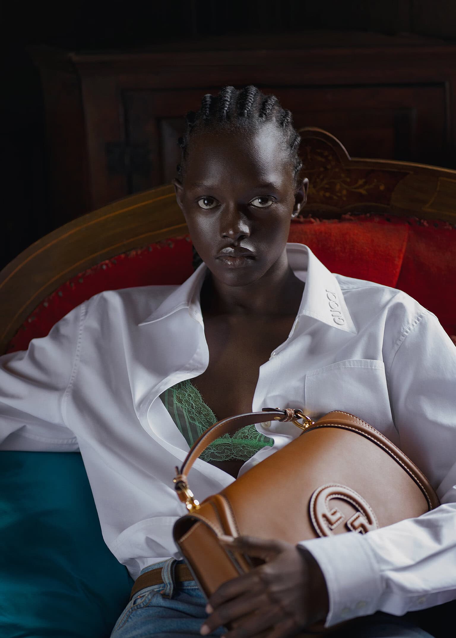 A Black woman seated on a red velvet sofa holding a tan leather bag while wearing a white shirt and green lace bra.