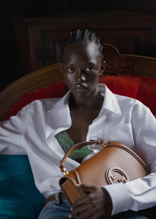 A Black woman seated on a red velvet sofa holding a tan leather bag while wearing a white shirt and green lace bra.