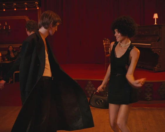 Woman in black dress dancing next to a man in a long dark coat inside a red-toned venue for Gucci We Will Always Have London campaign