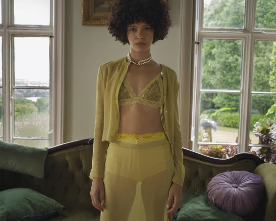 Model with dark curly hair wearing yellow sheer apparel and layered pearl necklace standing before a classic interior window