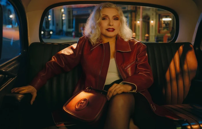 Debbie Harry seated in the back of a black taxi cab wearing a dark red leather jacket and holding a handbag.