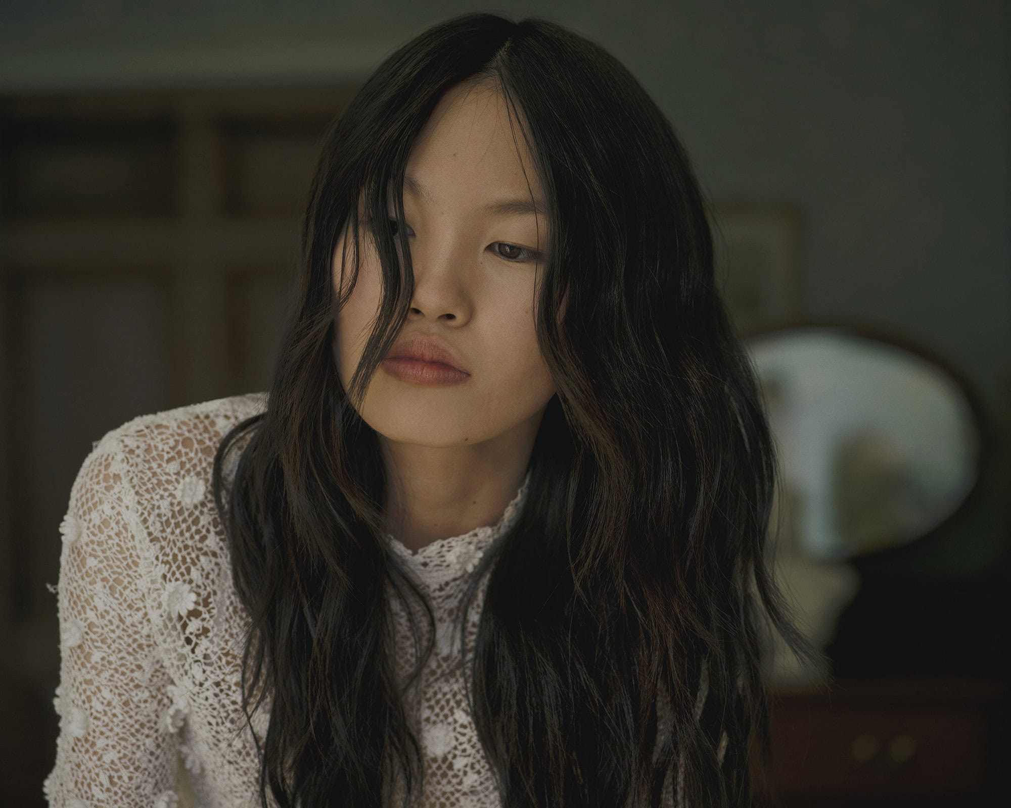Portrait of a woman with dark wavy hair wearing a white lace top in a dimly lit indoor setting.