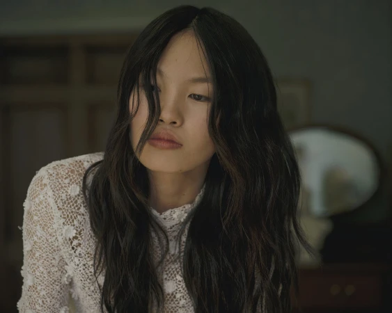 Portrait of a woman with dark wavy hair wearing a white lace top in a dimly lit indoor setting.