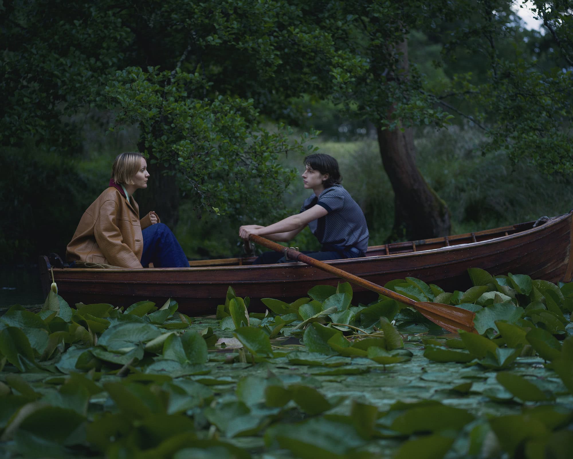 Two people seated in a wooden rowboat on a calm river surrounded by lush green lily pads and overhanging trees.