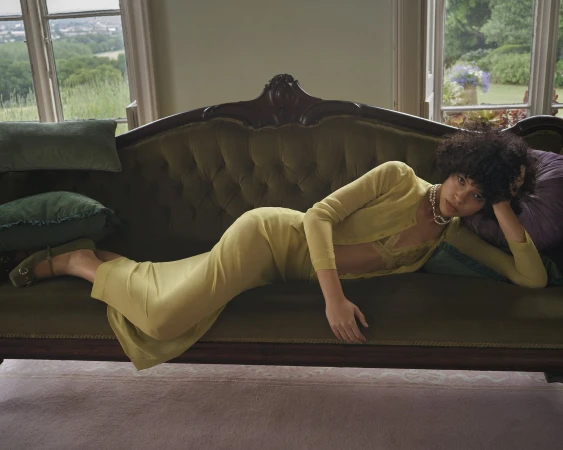 A woman reclining on a vintage green velvet sofa wearing a yellow cardigan and skirt set against a windowed interior.