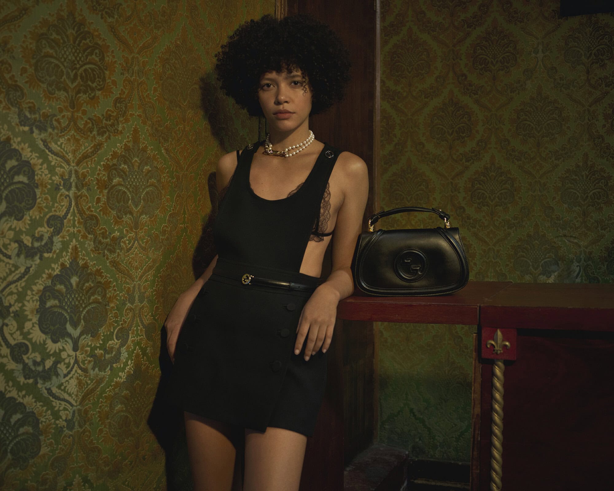 Model with dark curly hair wearing a black sleeveless dress and pearl necklace standing beside a black leather handbag against a patterned wallpaper interior