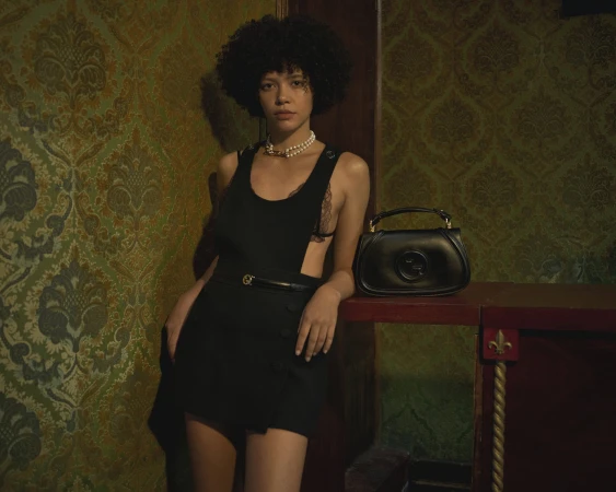 Model with dark curly hair wearing a black sleeveless dress and pearl necklace standing beside a black leather handbag against a patterned wallpaper interior
