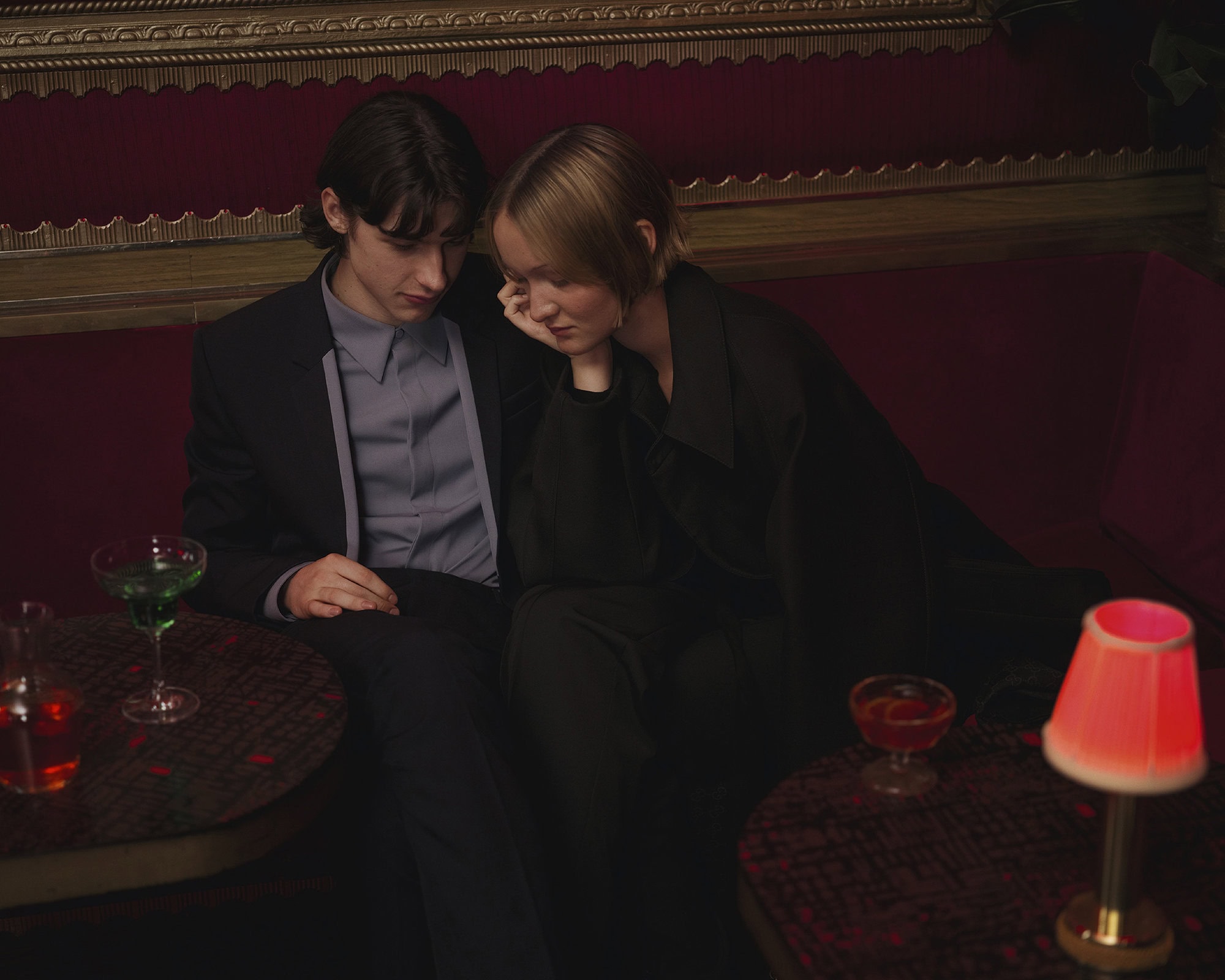 Two people seated on a red banquette in a dimly lit lounge with cocktails and a small lamp on a dark table.