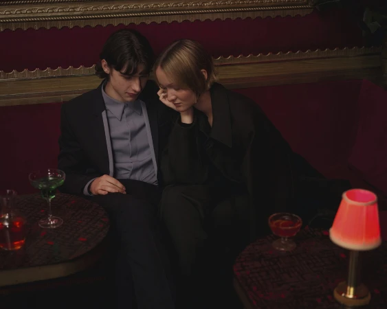 Two people seated on a red banquette in a dimly lit lounge with cocktails and a small lamp on a dark table.