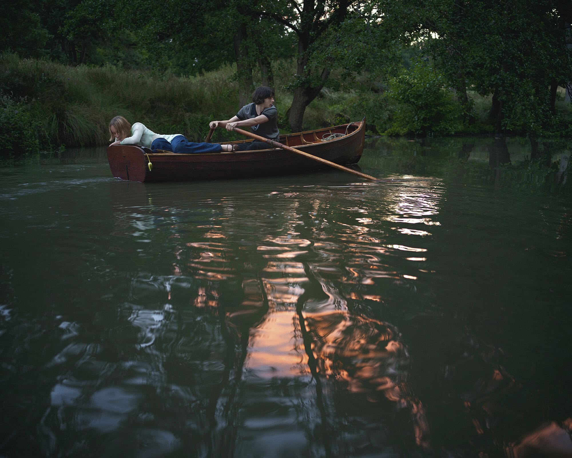 Two people in a wooden canoe on a calm river surrounded by lush green trees under overcast light