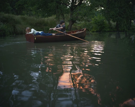 Two people in a wooden canoe on a calm river surrounded by lush green trees under overcast light