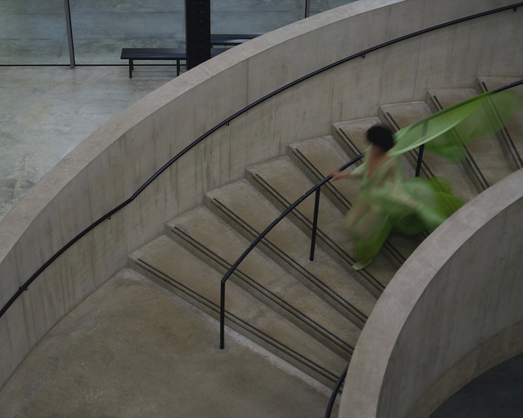 A person in a flowing green dress walking down a curved concrete staircase with a black handrail.