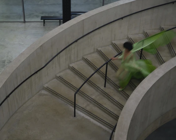 A person in a flowing green dress walking down a curved concrete staircase with a black handrail.