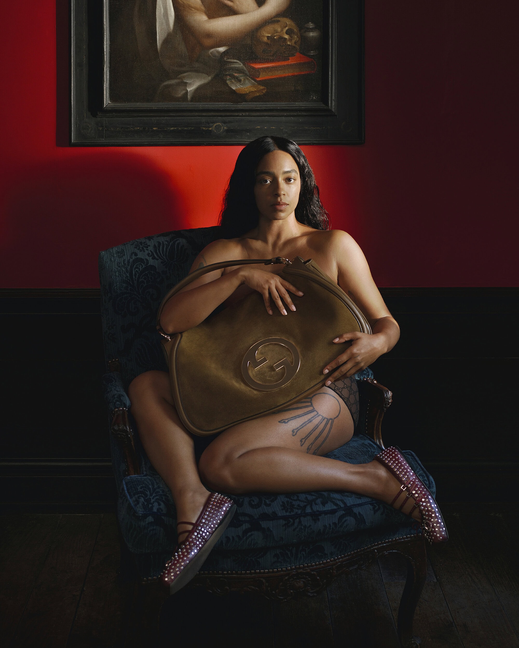 A person seated in a patterned armchair holding a large olive green handbag in front of a red wall with a framed painting.