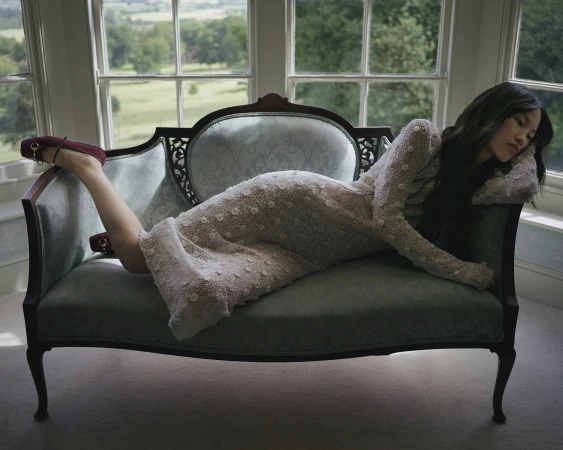 Woman in a long textured white dress lounging on an antique settee before large windows overlooking green landscape.