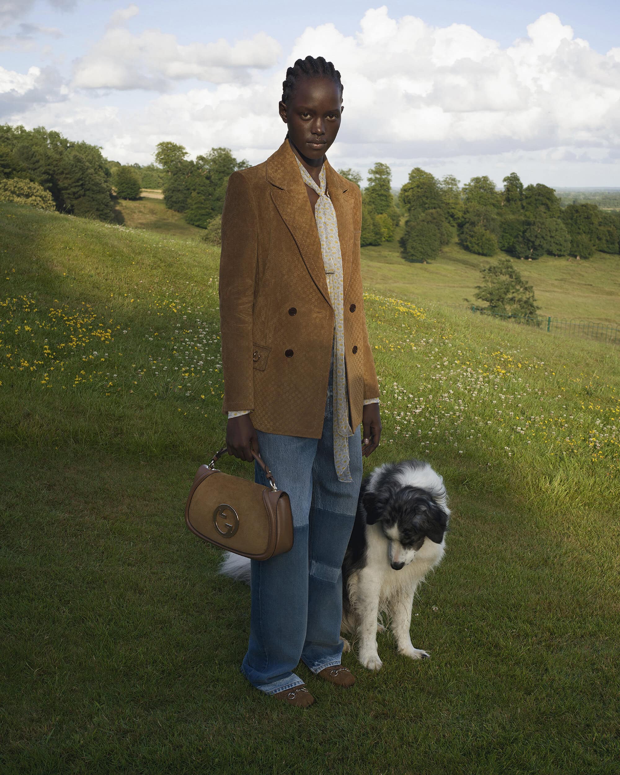 Model wearing a brown blazer and jeans standing on a grassy hill next to a dog with a handbag.