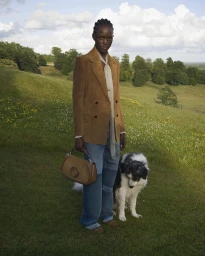 Model wearing a brown blazer and jeans standing on a grassy hill with a dog for Gucci