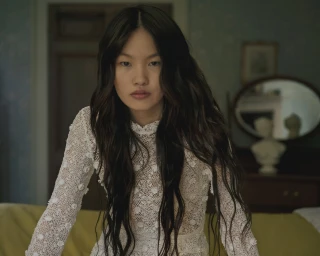 Model with long dark wavy hair wearing a white lace dress in a domestic room